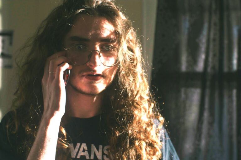 Still from I Can Be Killed: a young man with long curly hair and round glasses holding a phone to his ear