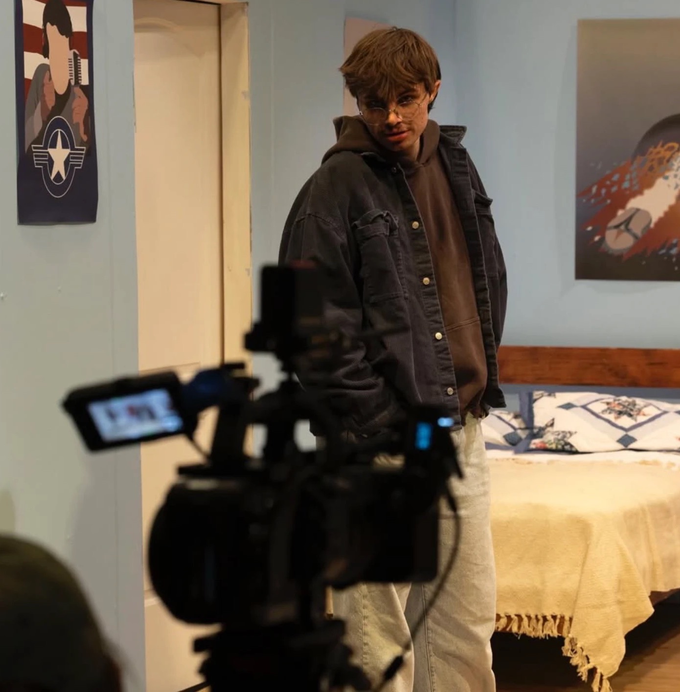 Behind the scenes on a film set: a person in round glasses and a dark jacket stands in a bedroom set behind a camera